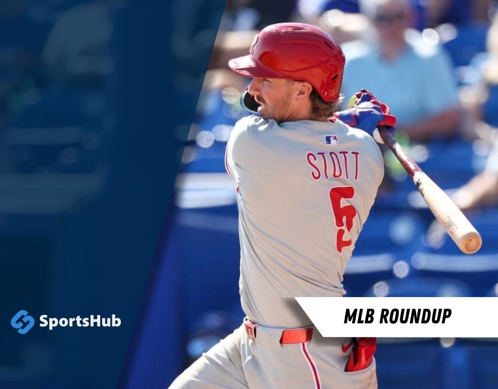 A baseball player in a light grey uniform with red accents prepares to bat during a game, with spectators in the background.