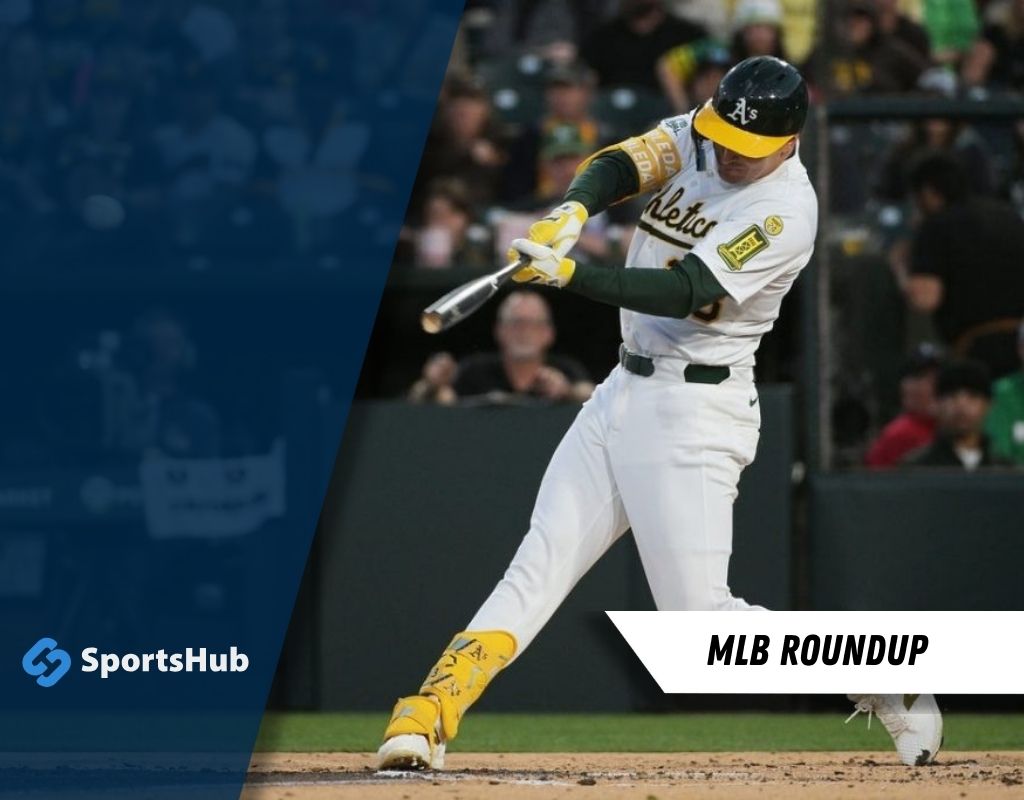 A baseball player in an Oakland Athletics uniform swings a bat, with fans and stadium seating blurred in the background.