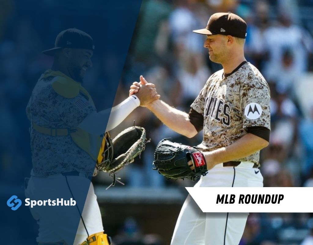 Two baseball players in matching uniforms shake hands, with one holding a glove and the other a baseball cap in a sunny stadium.