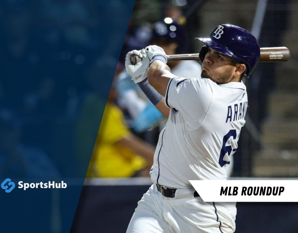 A player in a white baseball jersey with "ARAUJO" on the back swings a bat during a night game, with spectators blurred in the background.