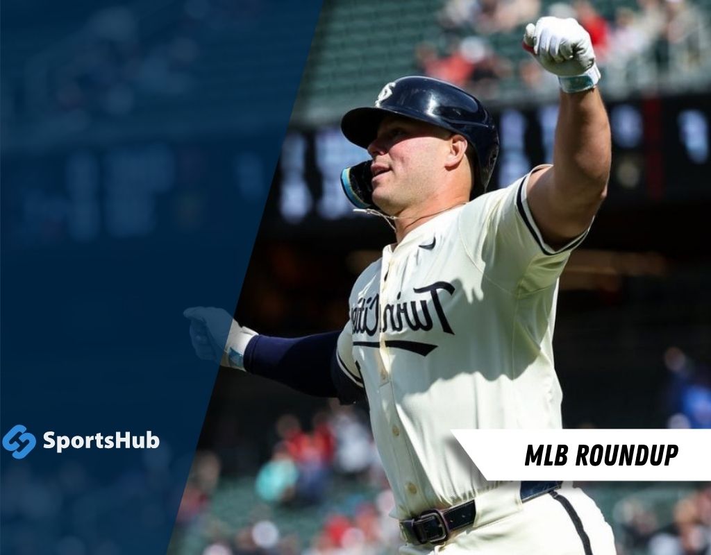 A baseball player in a white uniform celebrates enthusiastically, pumping his fist, with a stadium full of fans in the background.