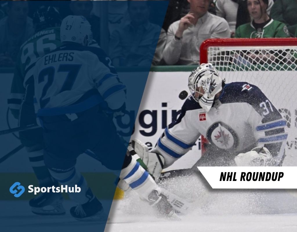 A goalie in a white and blue uniform dives to stop a puck during an NHL game while players skate in the background. SportsHub branding visible.